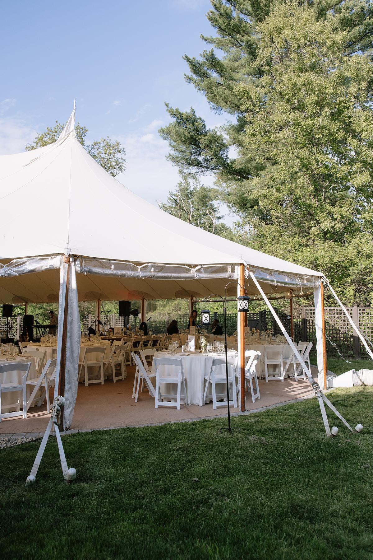 Large white event tent on grass, surrounded by trees, with tables and chairs inside.