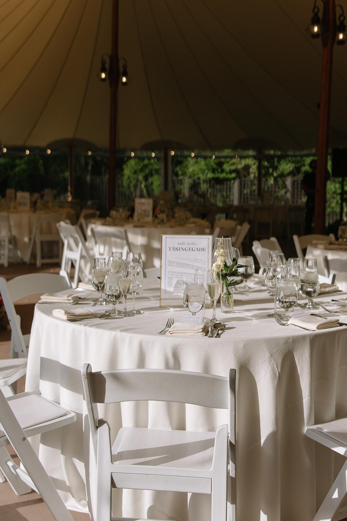 Elegant table setting under a canopy, with white chairs and floral centerpieces.