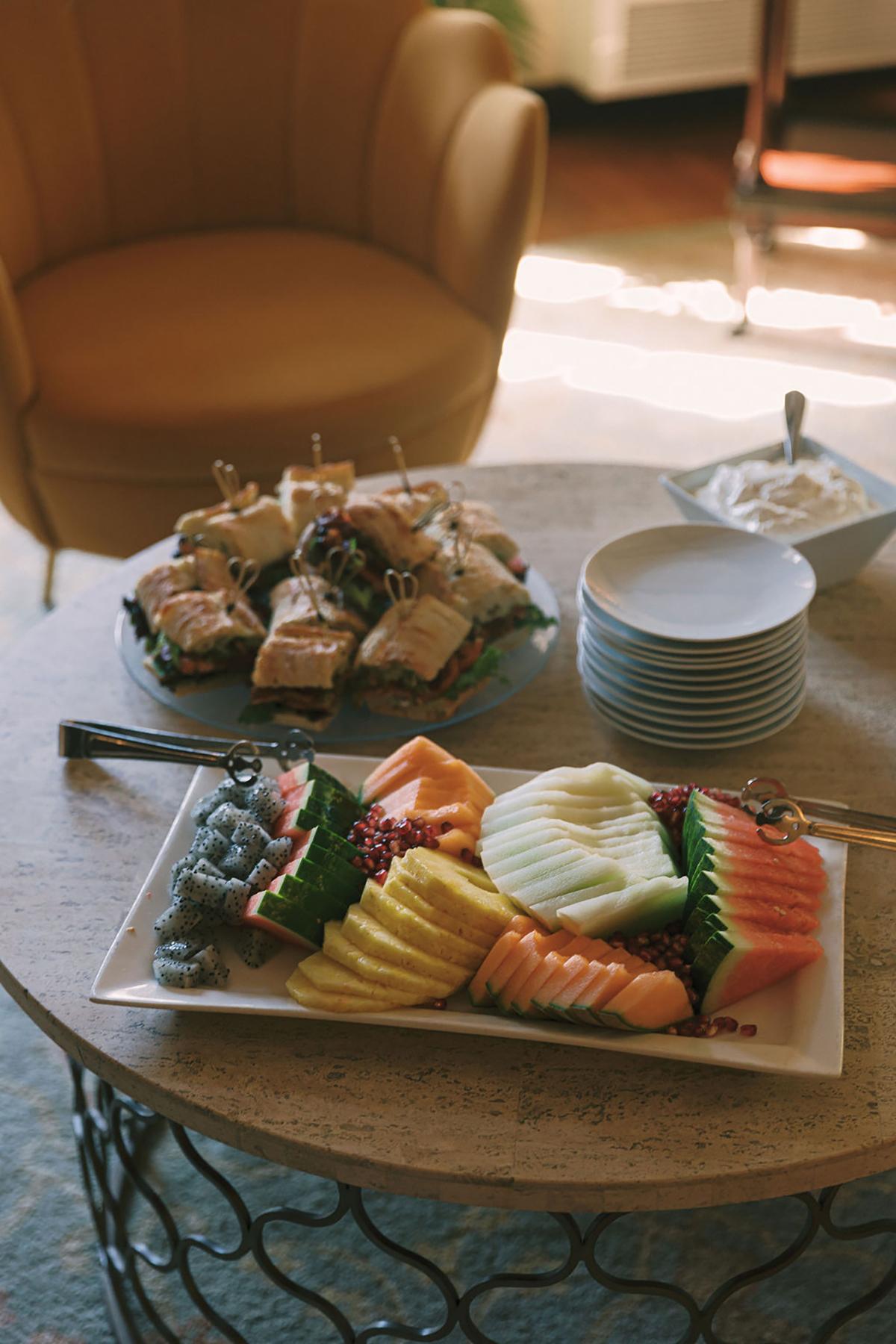 Fruit platter and sandwiches on a round table near a yellow armchair.