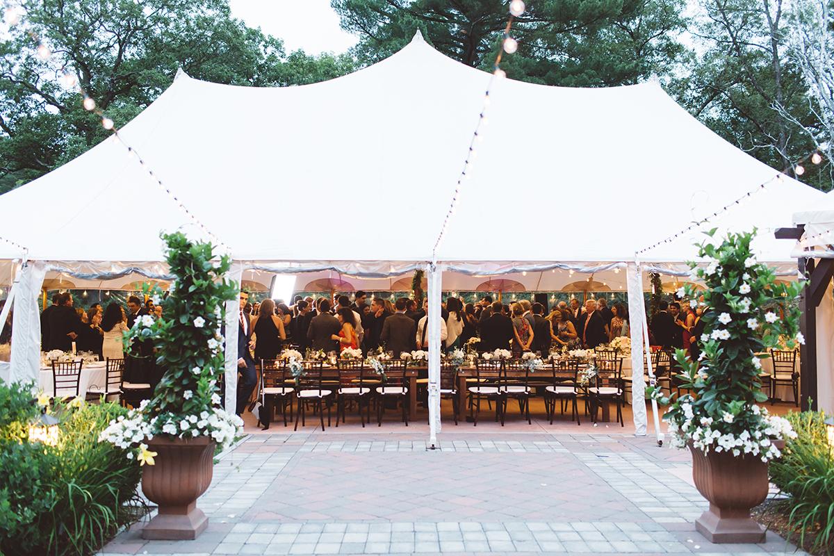 White event tent with people dining inside, surrounded by greenery.