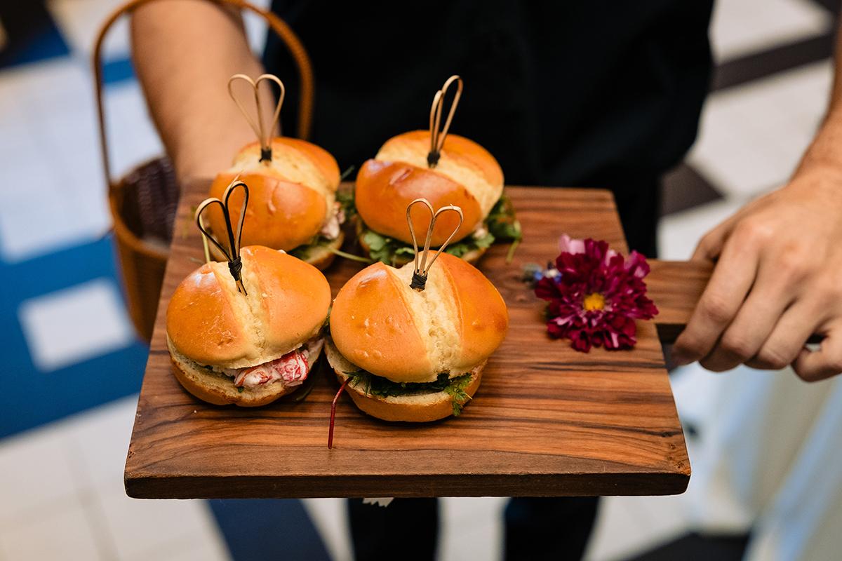 Four mini burgers on a wooden tray with a flower garnish.