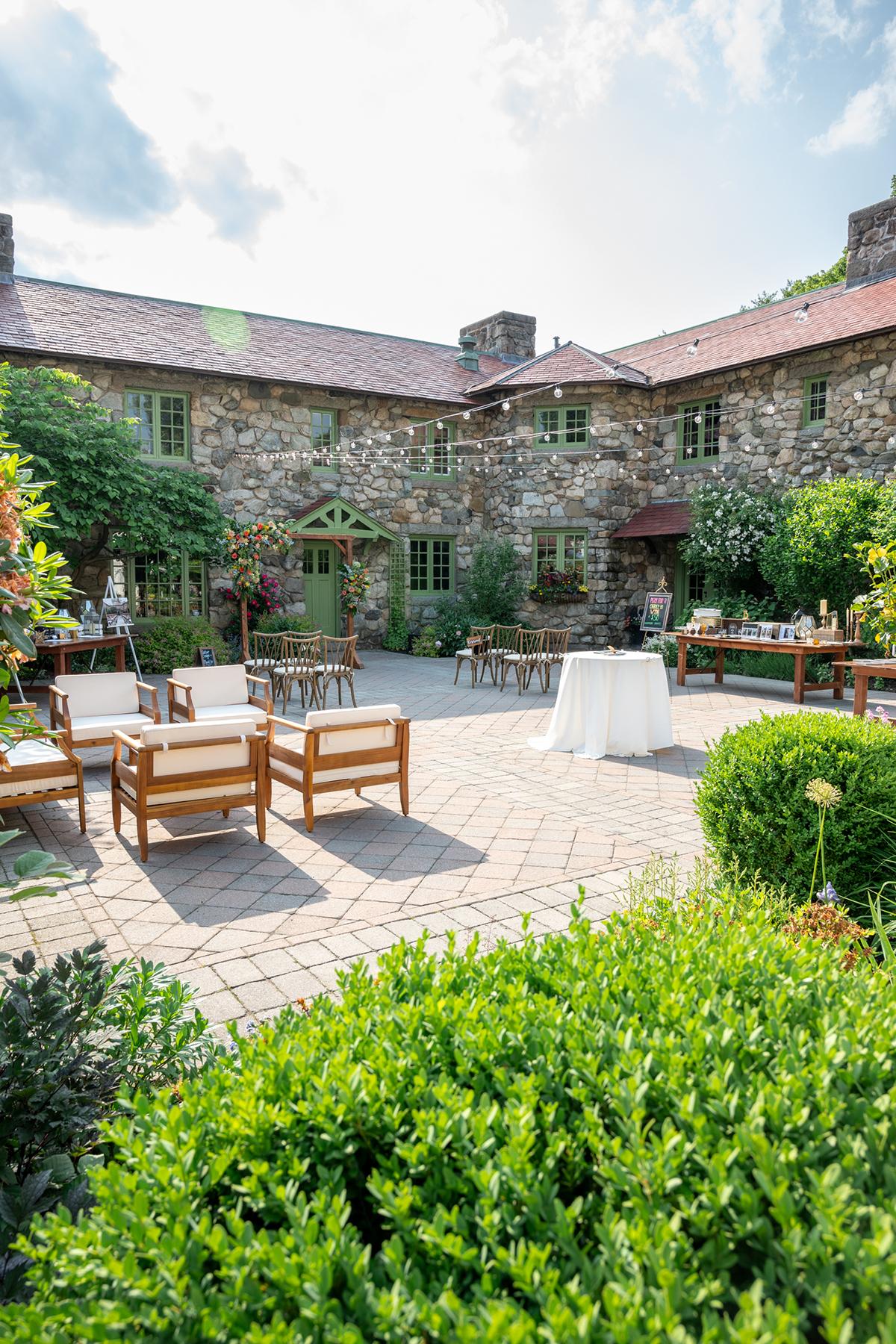 Sunny courtyard with stone building, patio furniture, and lush greenery.