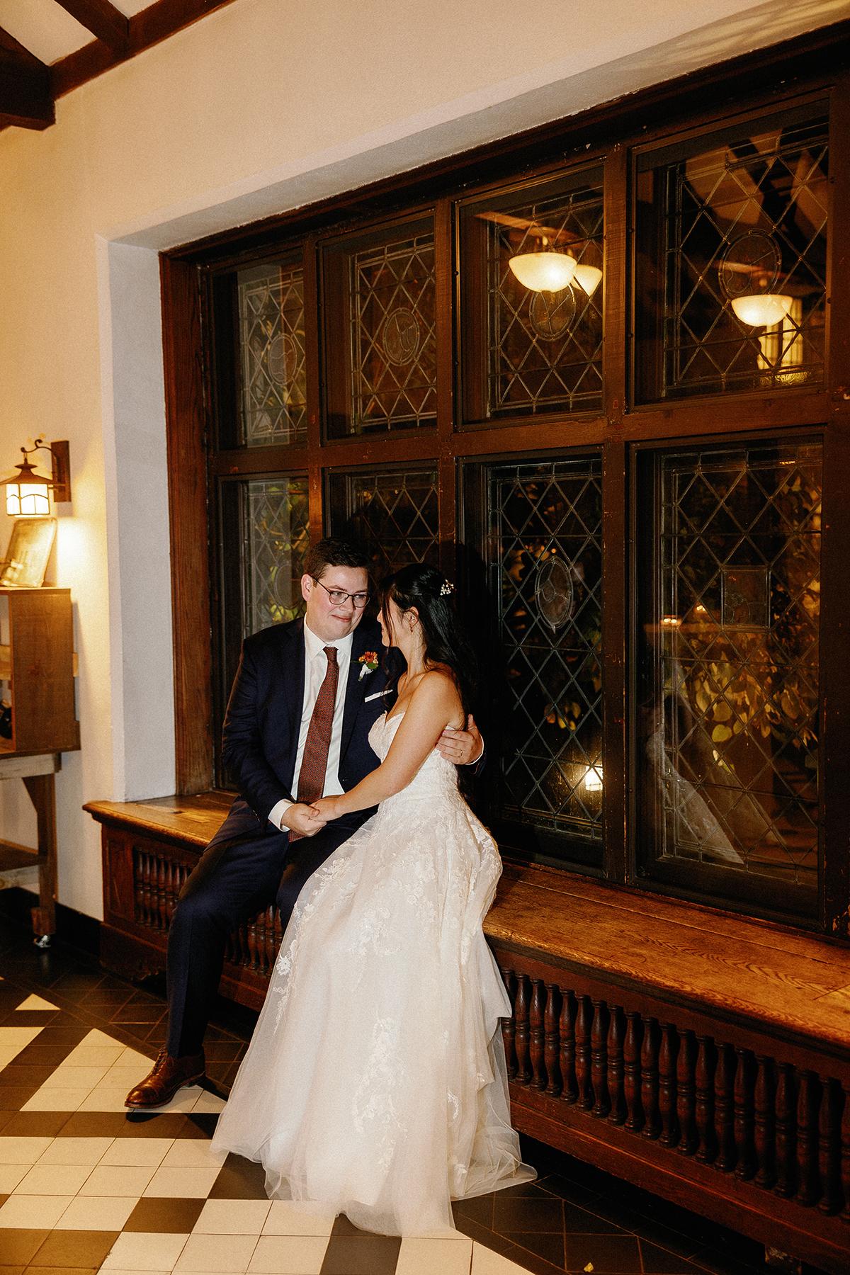 Bride and groom sitting by a large window at night, smiling at each other.