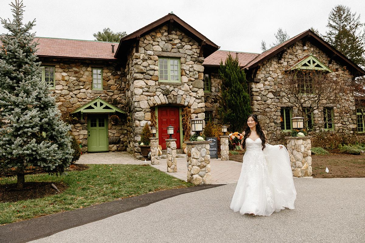 Bride holding bouquet, walking in front of stone house with green doors.