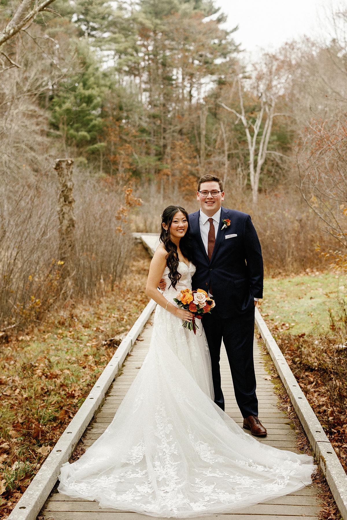 Bride and groom smiling on a wooden path surrounded by autumn foliage.