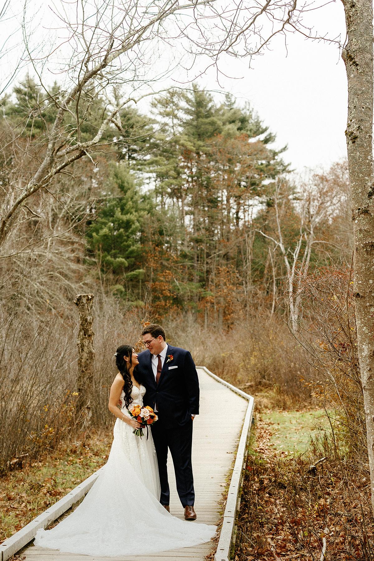 Bride and groom standing on a wooden path in a forest, embracing tenderly.