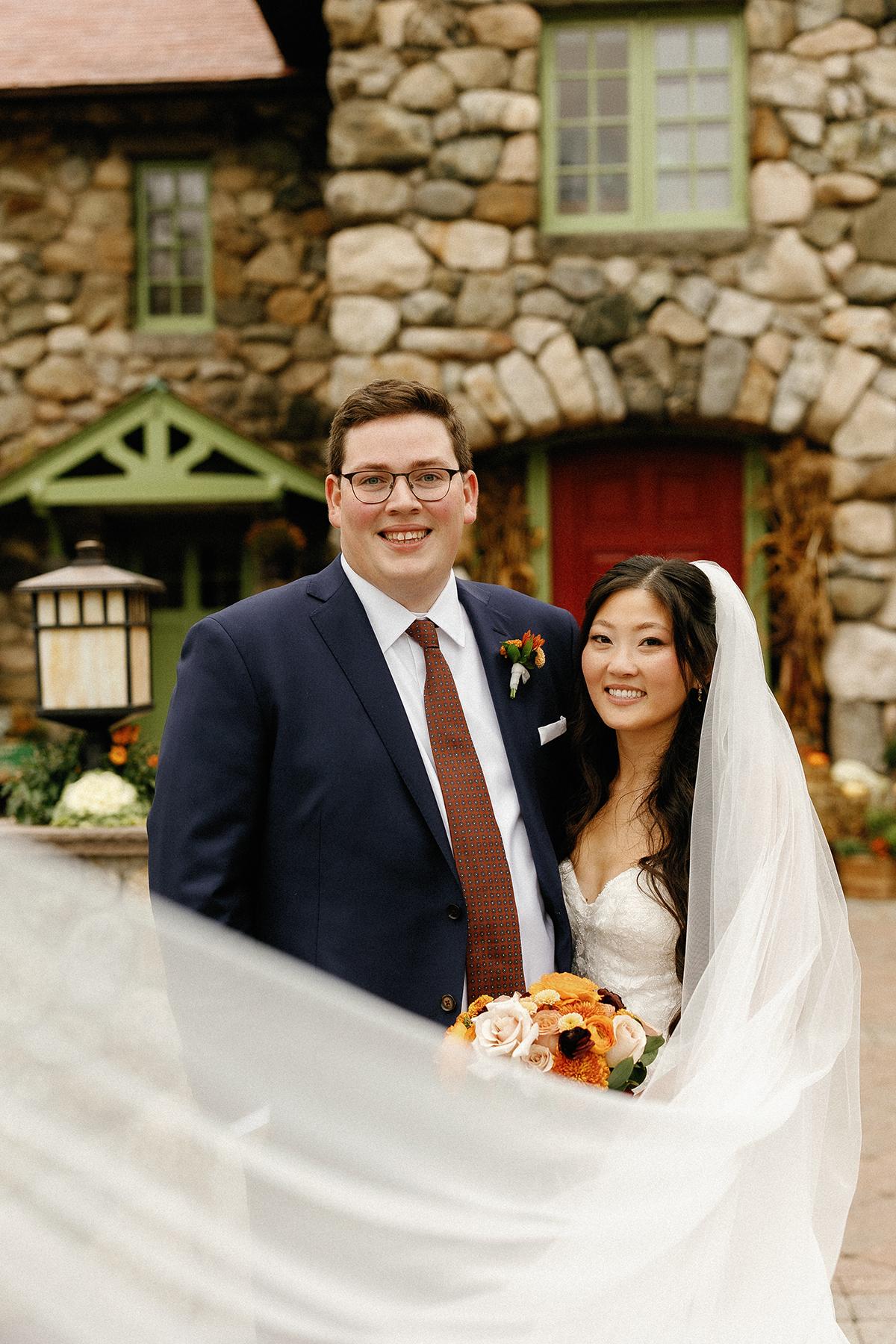 Bride and groom smiling in front of a stone building, veil flowing.
