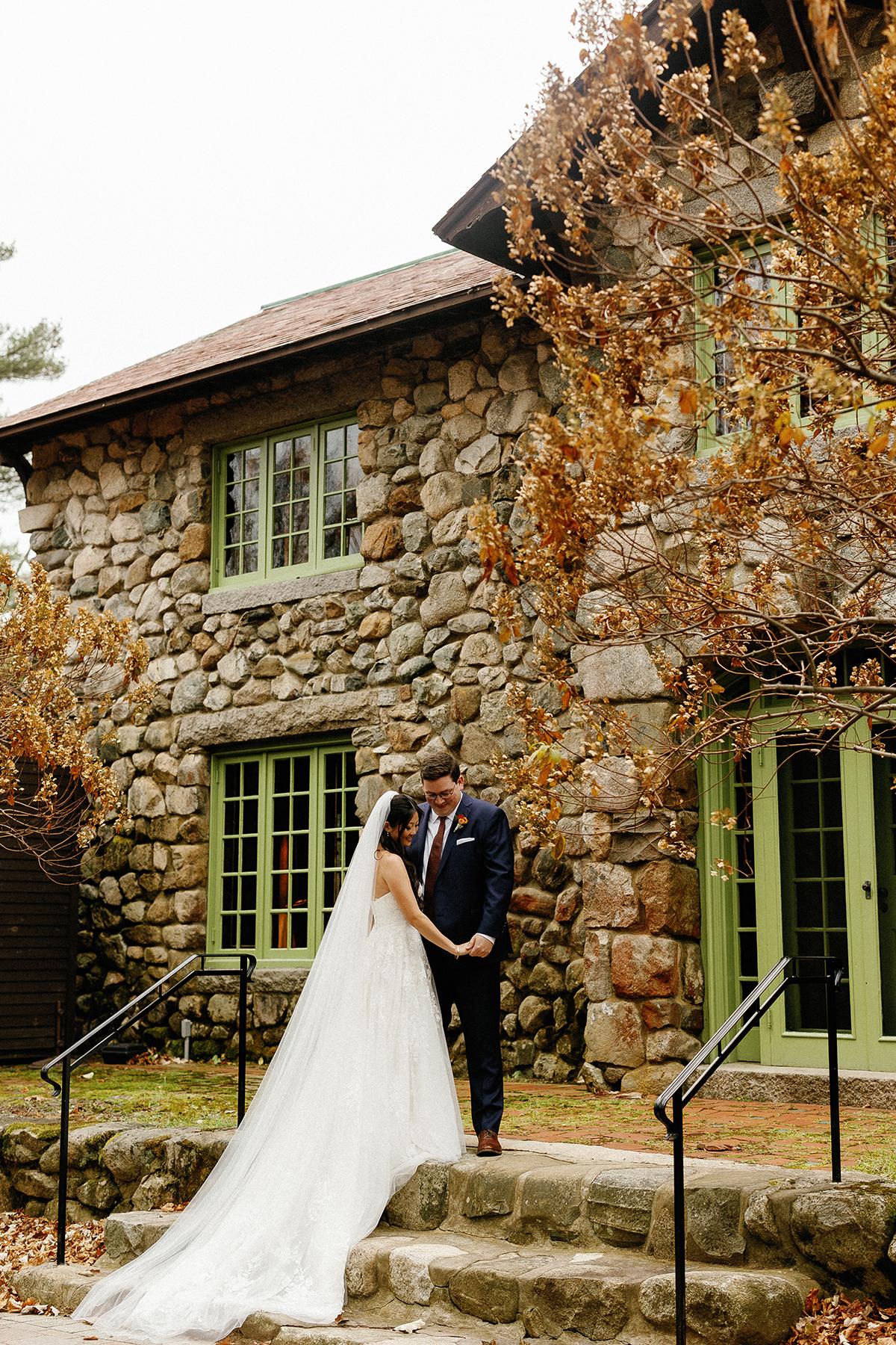 Bride and groom embrace outside a stone house with green windows.