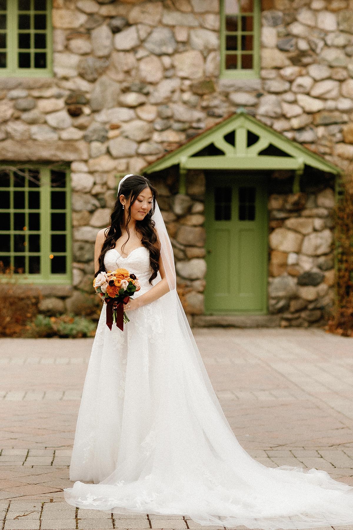 Bride in white dress holding bouquet, standing by stone building with green door.