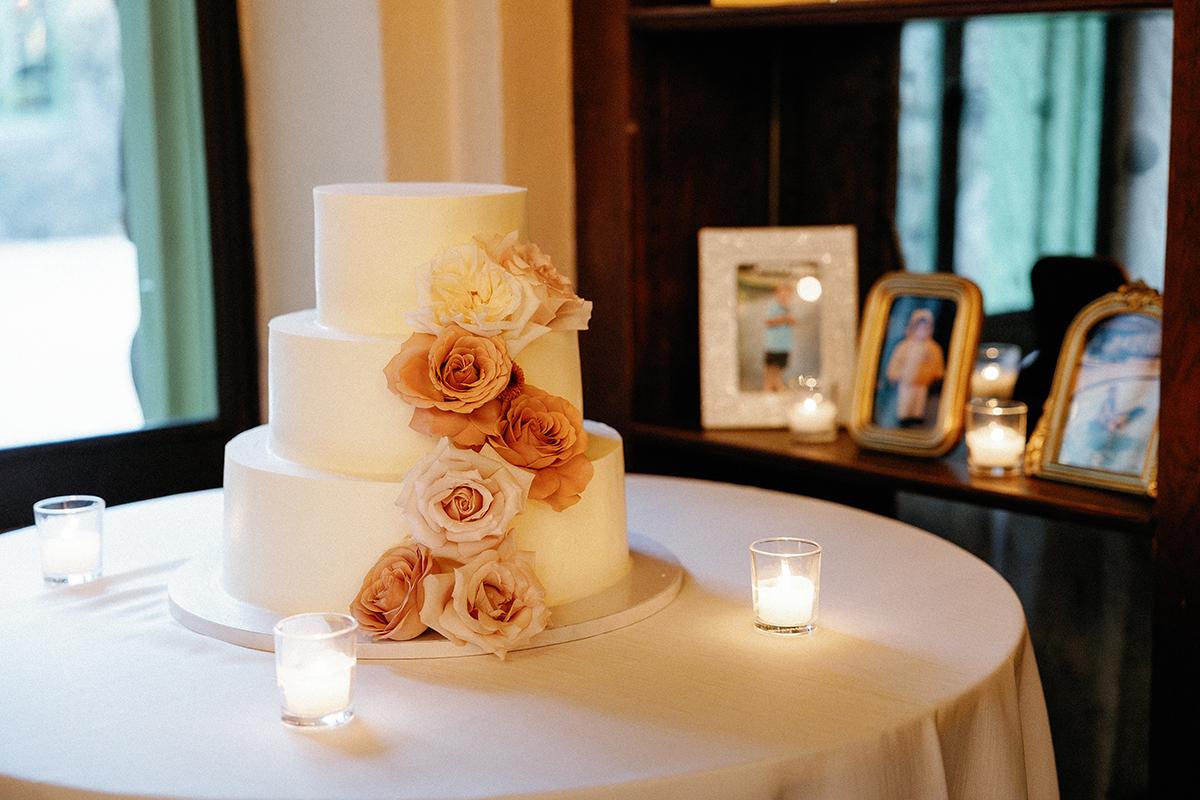 Three-tiered white wedding cake with pink roses, surrounded by candles and framed photos.