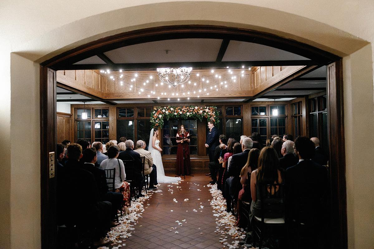 Wedding ceremony under arches and string lights, guests seated on either side.