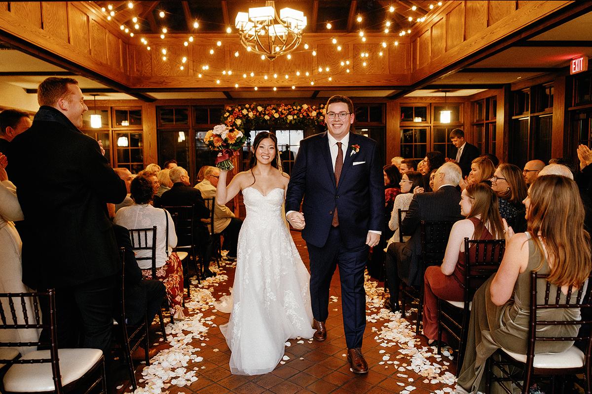 Bride and groom walking down aisle, smiling, under string lights at indoor wedding ceremony.