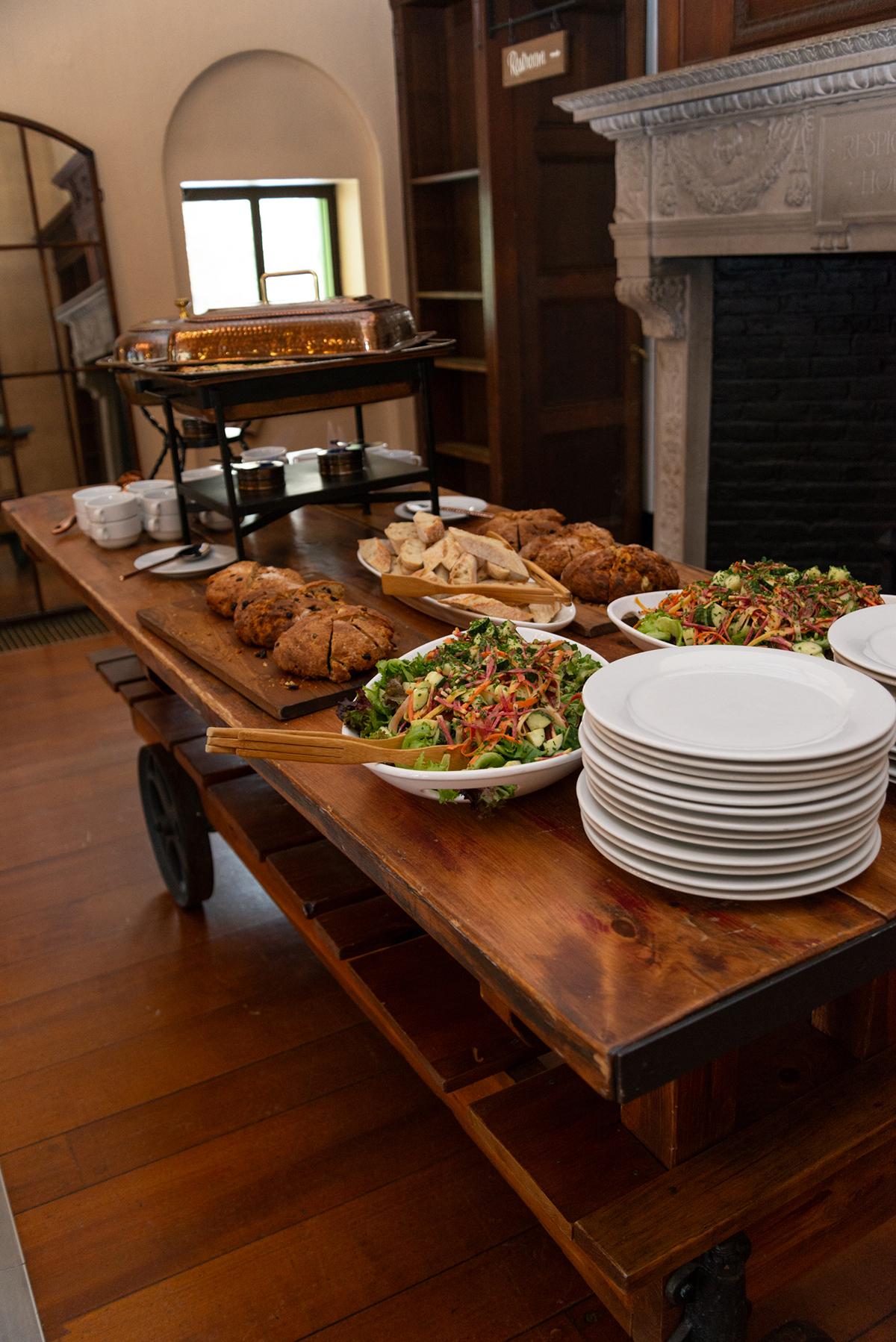 Buffet table with salads, bread, and stacked plates in a cozy room.