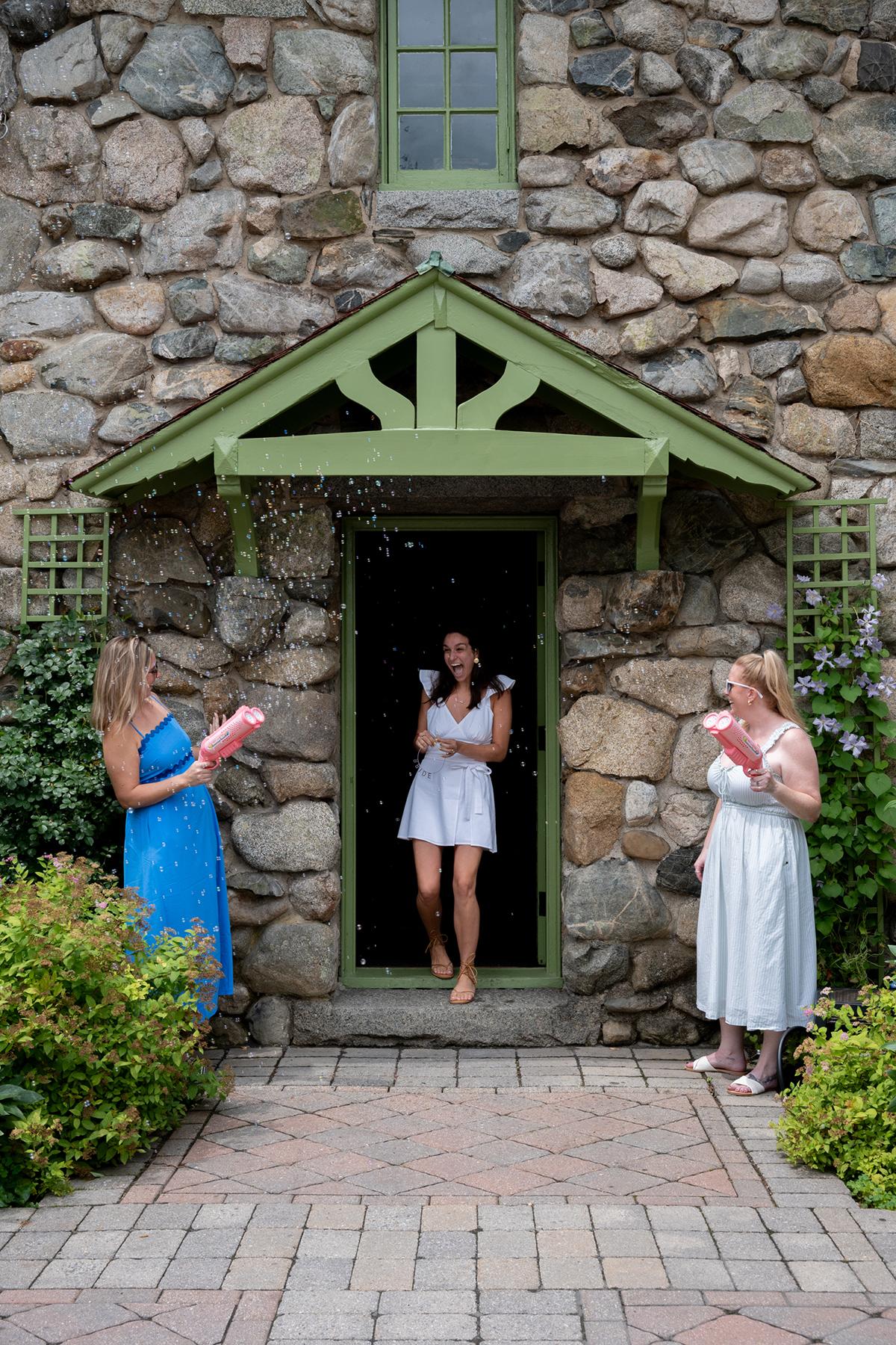 A woman in a white dress stands in a doorway, smiling at two women holding pink boxes.