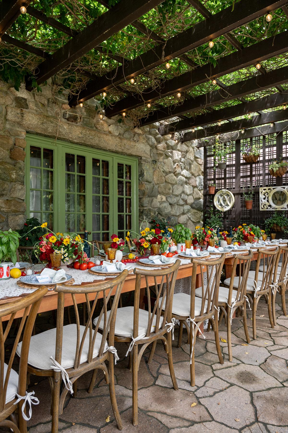 Outdoor dining table with colorful flowers under a pergola, against a stone wall.