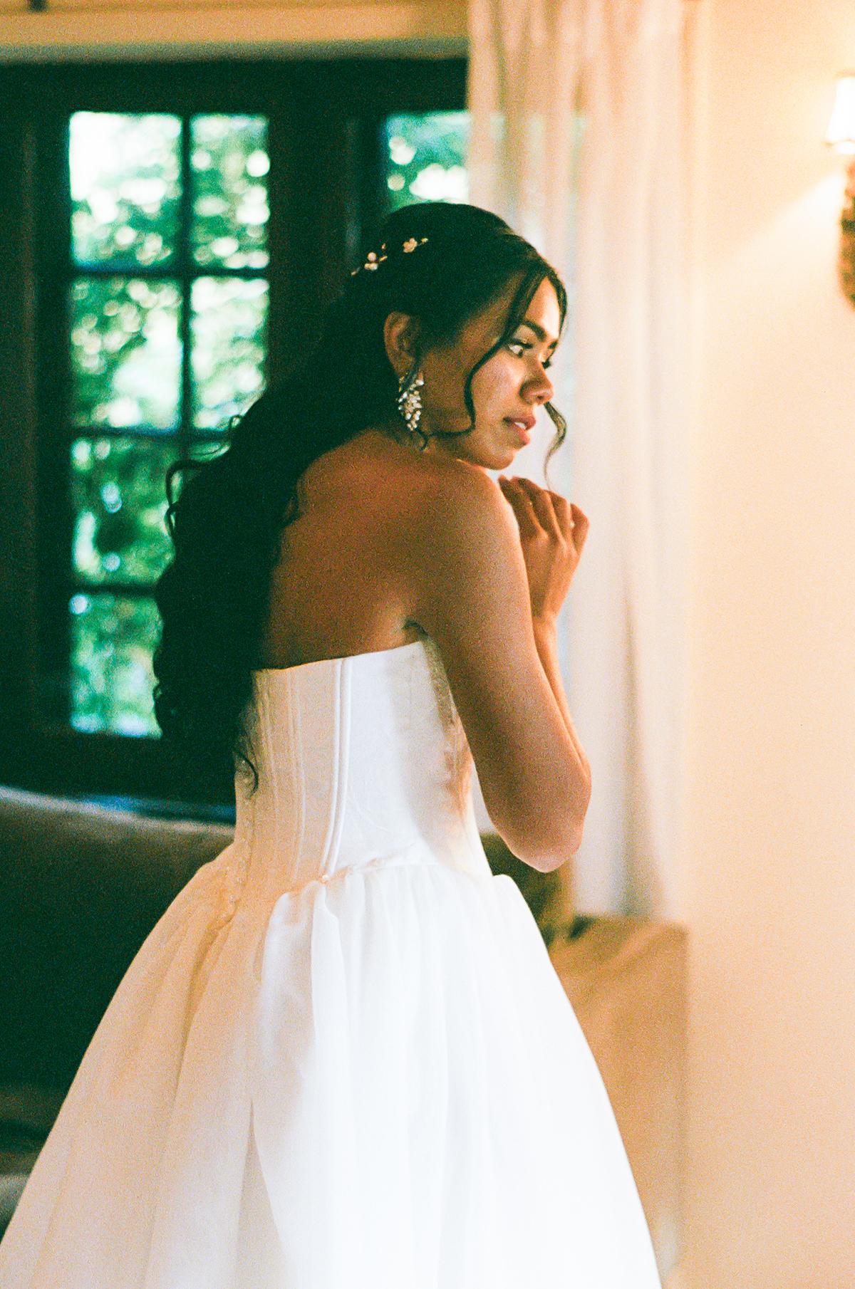 Bride in a white gown stands by a window, softly lit, looking thoughtful.