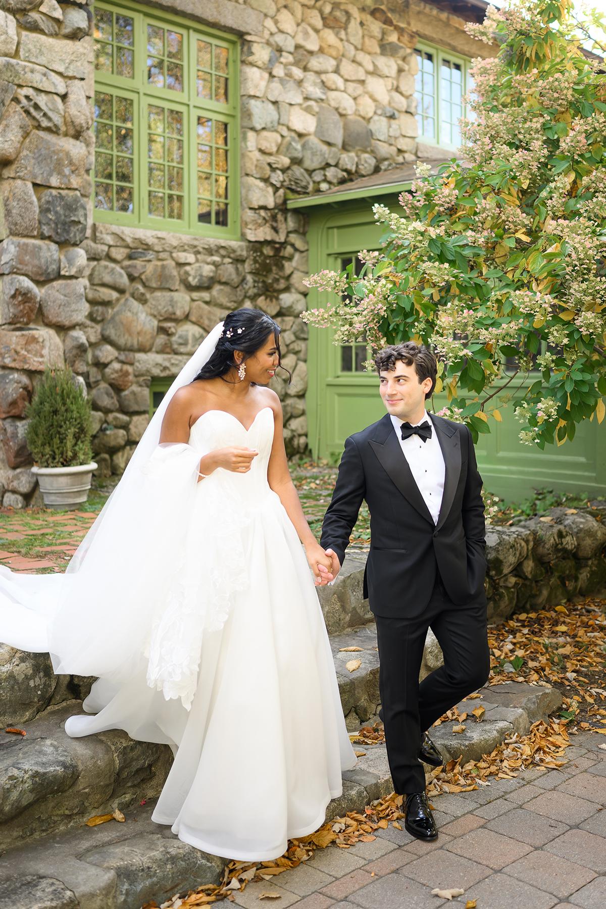 Bride and groom holding hands, walking by a stone building in autumn.