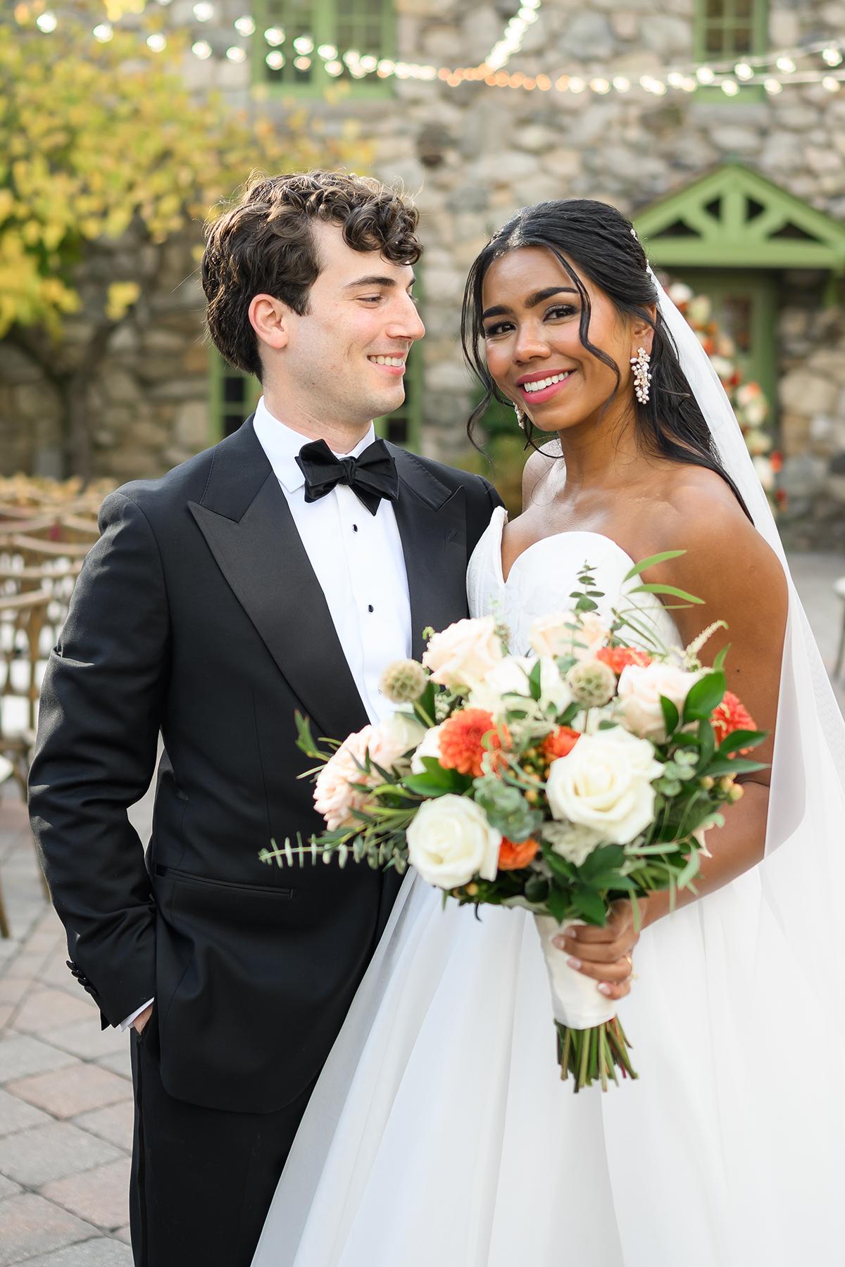 Bride and groom smiling, she holds a bouquet of white and orange flowers.