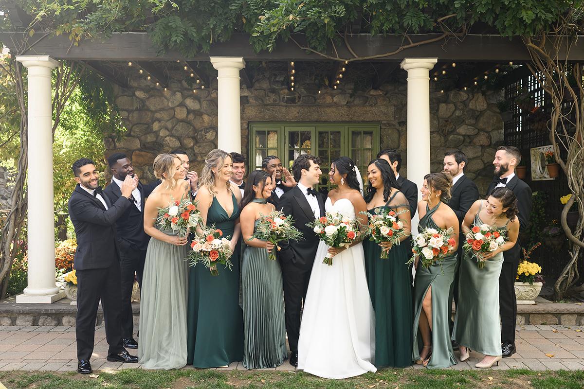 Wedding party smiling under a pergola, bridesmaids in green dresses, groomsmen in black suits.