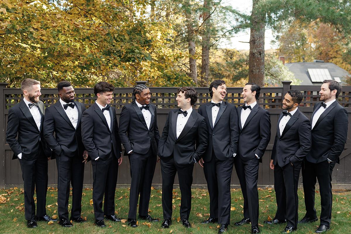 Groomsmen in tuxedos stand together outdoors on a fall day.