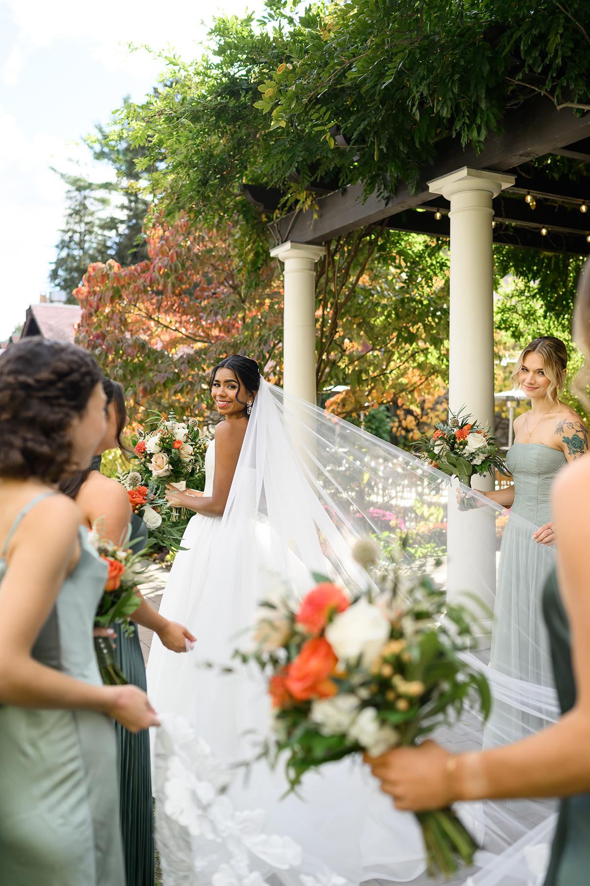 Bride in white dress with veil and bridesmaids holding flowers outside.