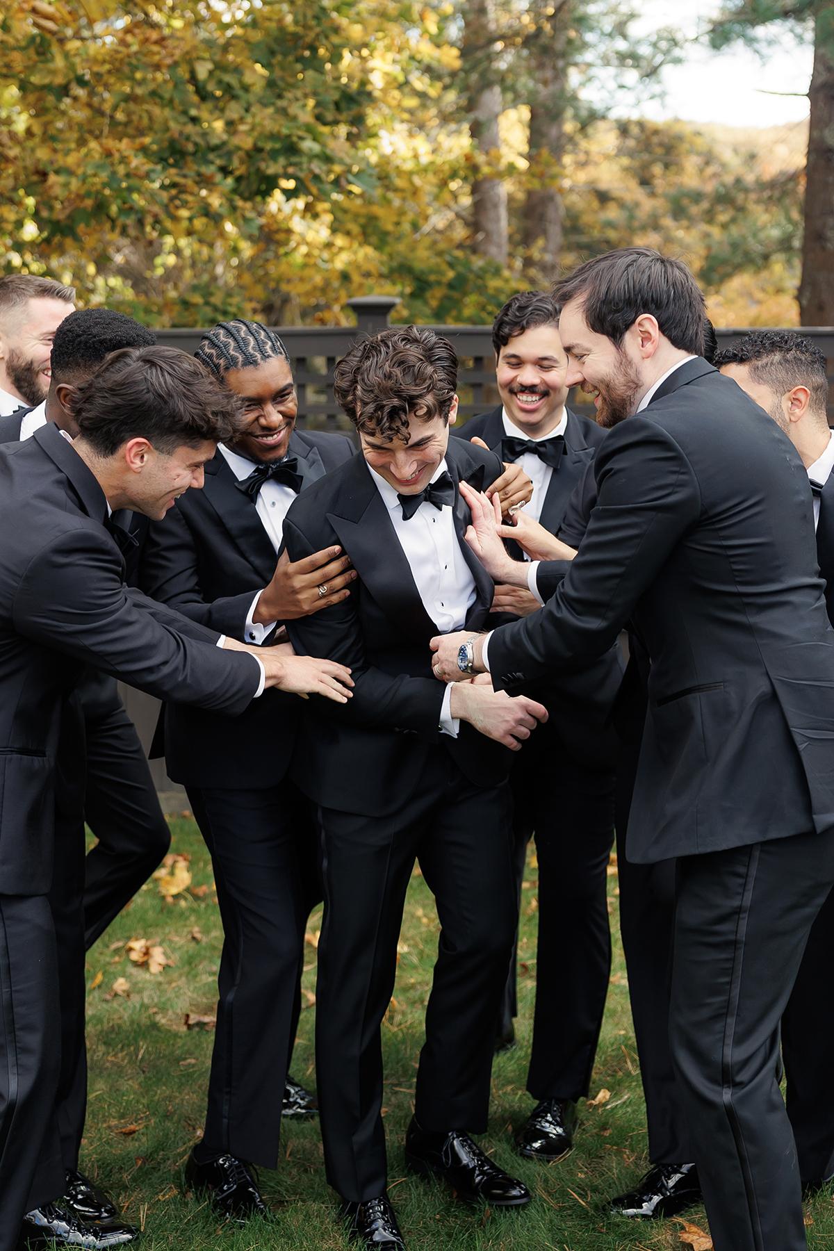 Groom and groomsmen in suits laugh together outdoors.