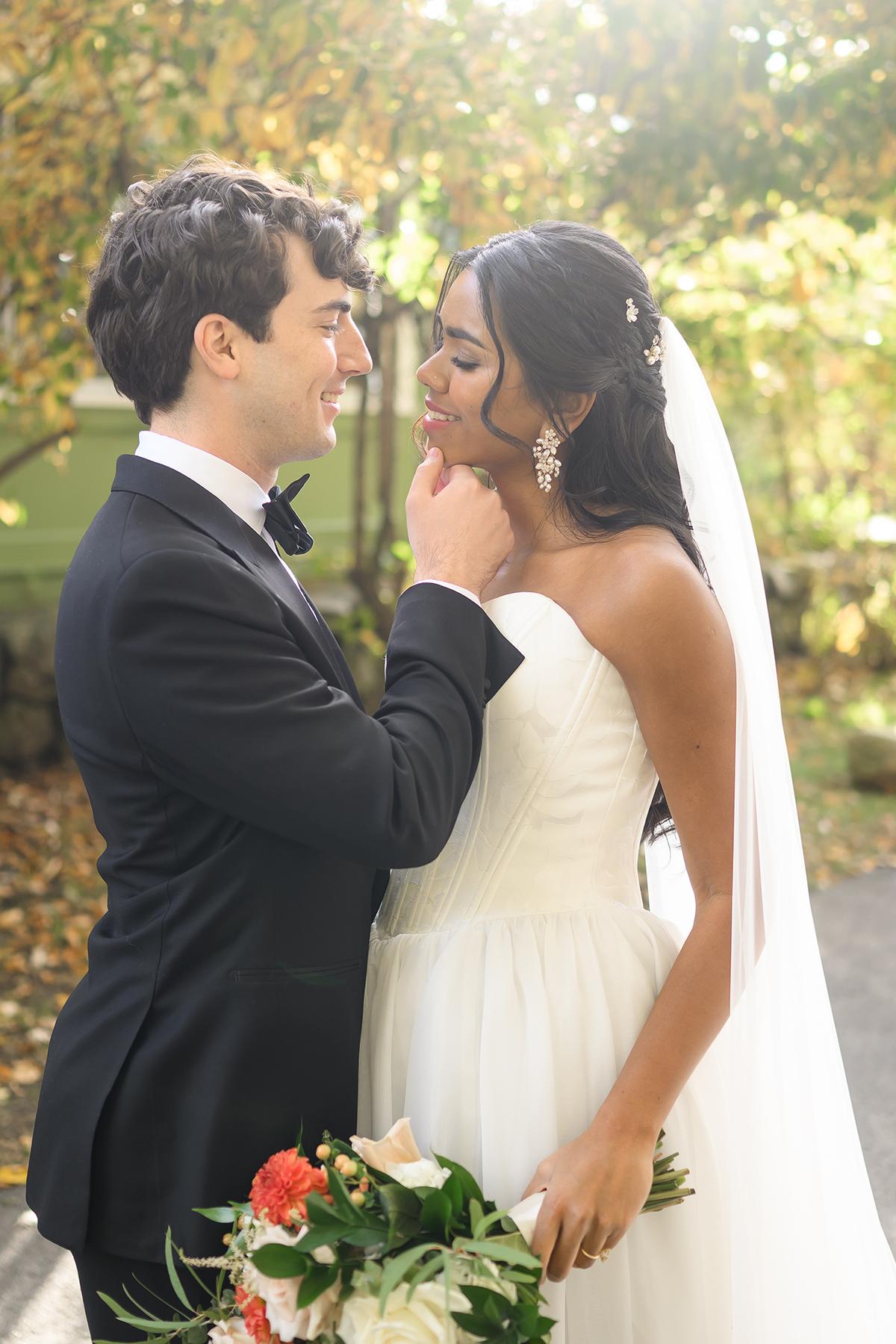 Bride and groom smiling at each other, holding a bouquet, in a sunlit garden.