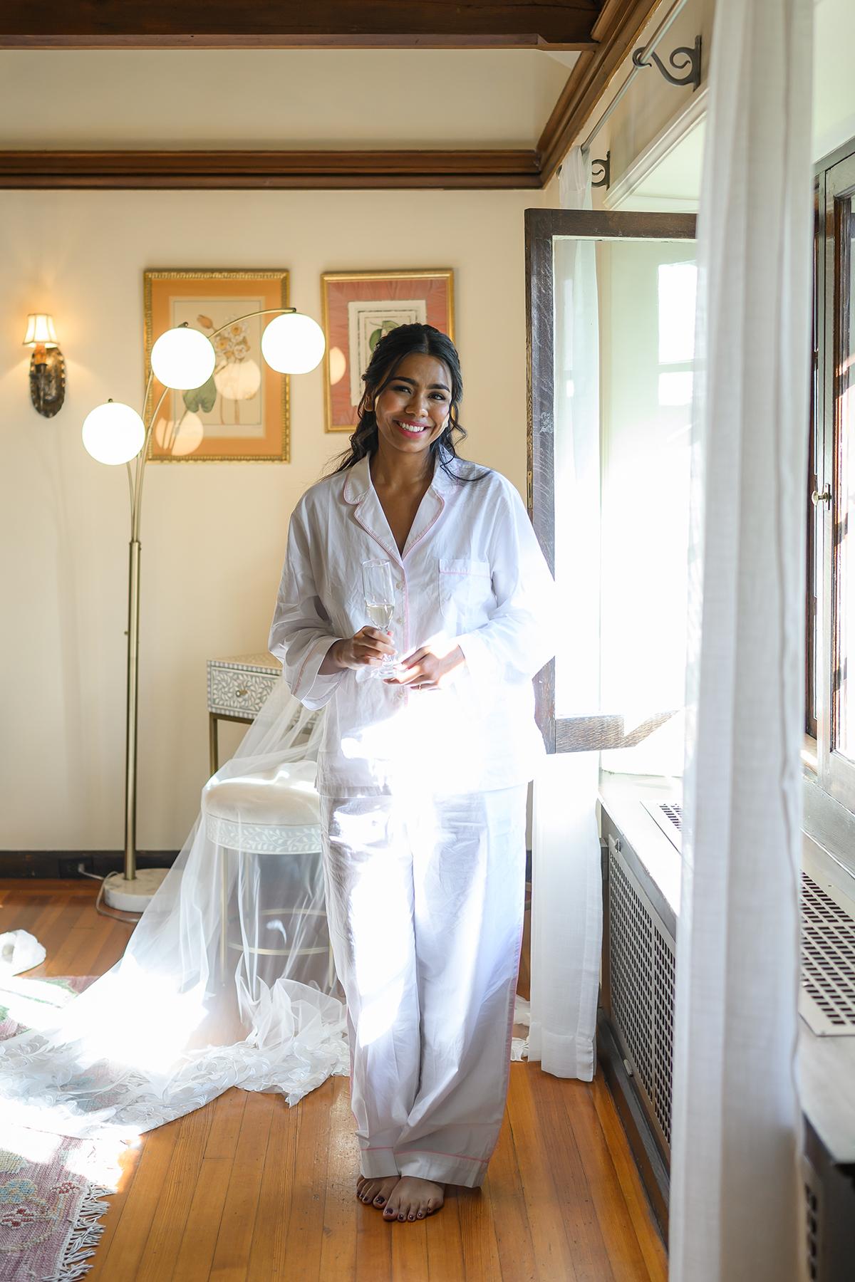 Woman in white pajamas stands by a sunlit window, smiling.