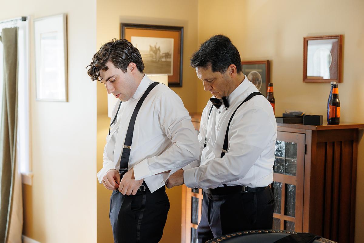 Two men in formal wear adjusting suspenders in a warmly lit room.