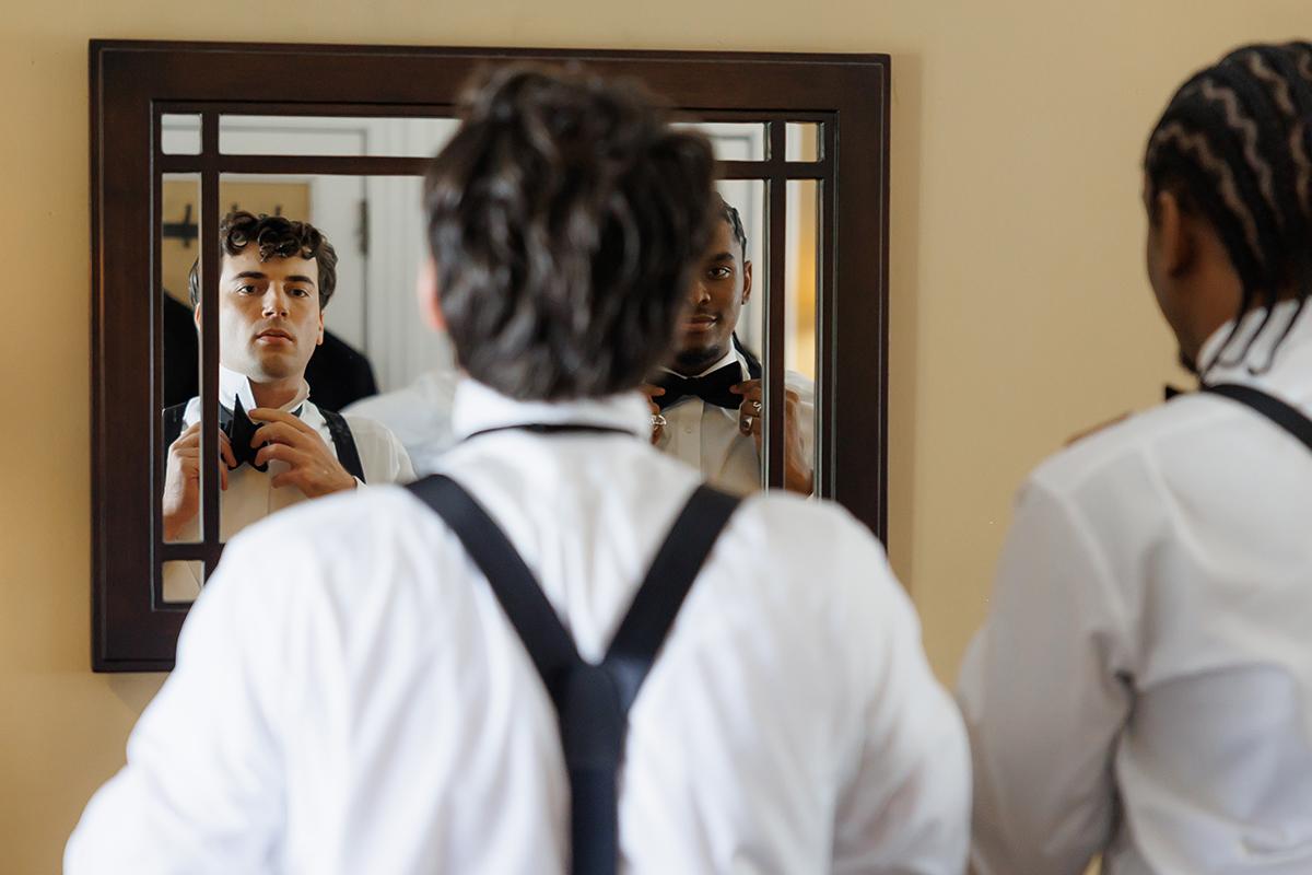 Two people wearing formal attire adjusting bow ties in a mirror.