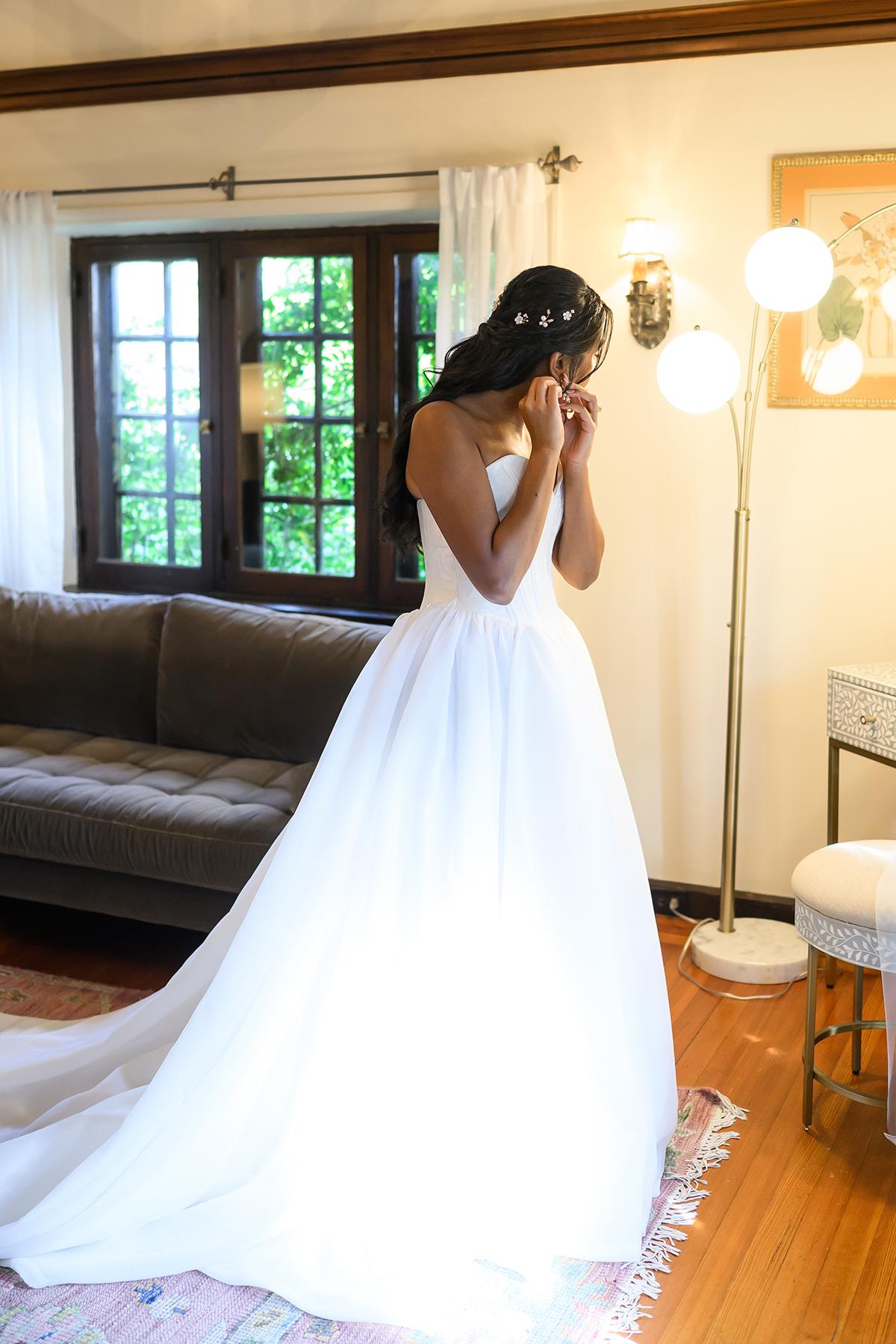 Bride in white gown standing in a warmly lit room, near a couch and floor lamp.