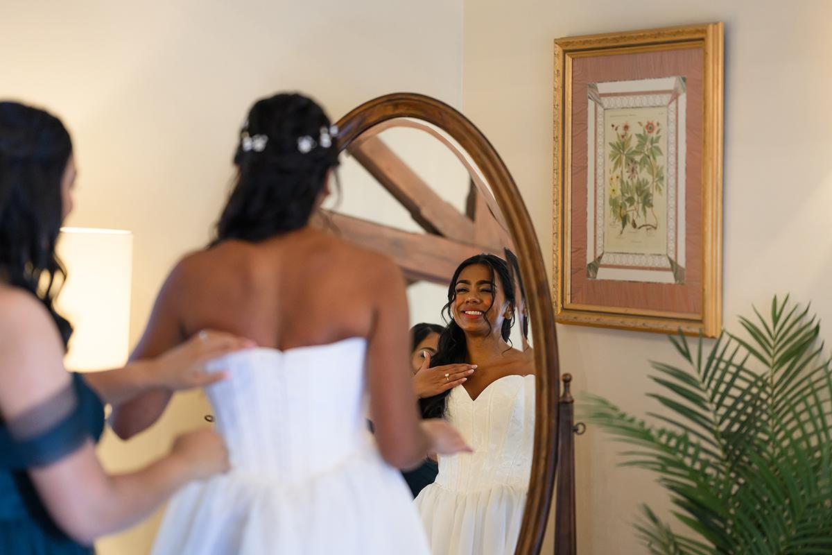 Bride smiling in mirror, adjusting dress with help in warmly lit room.