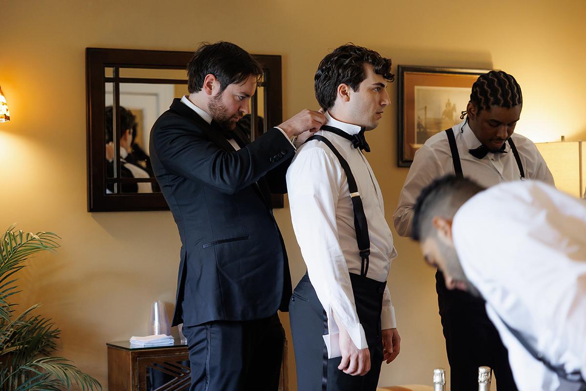 Men in suits adjusting their bow ties in a warmly lit room.