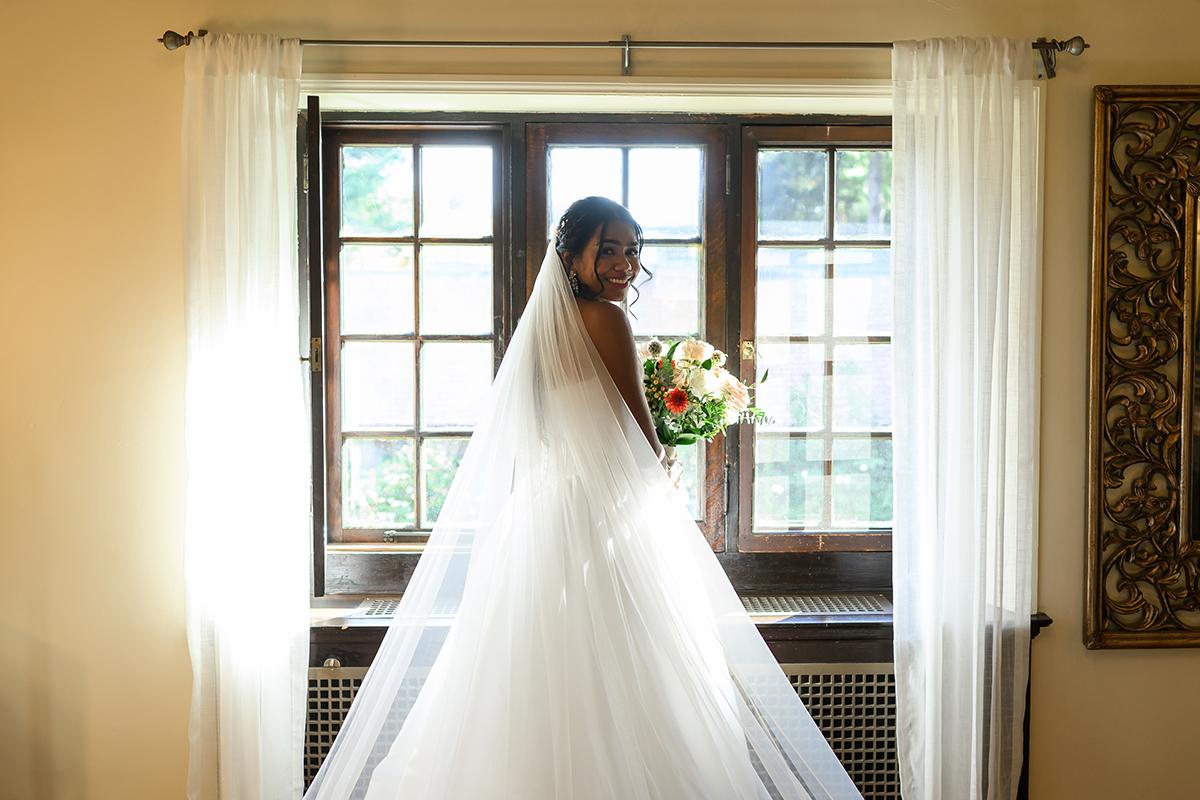 Bride in white gown holds bouquet, stands by window with sheer curtains.