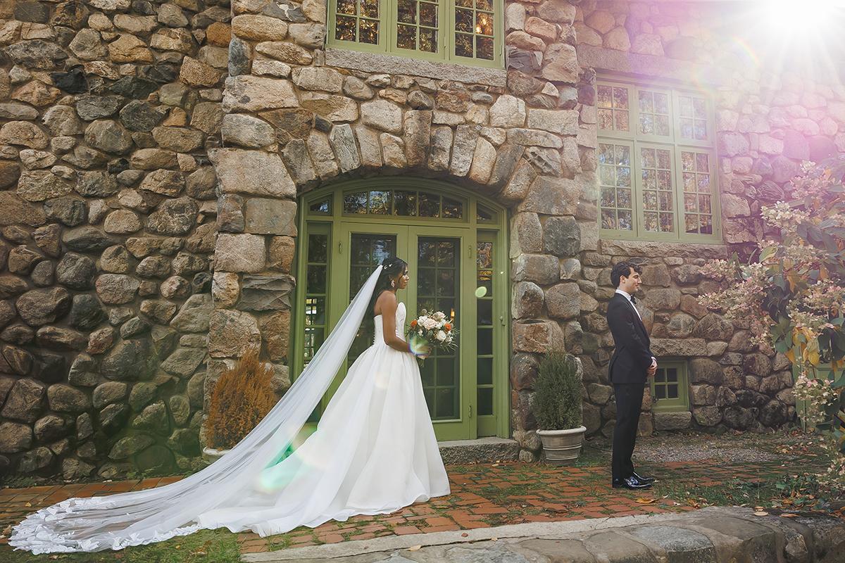 Bride and groom stand outside a stone building, sunlight streaming in.