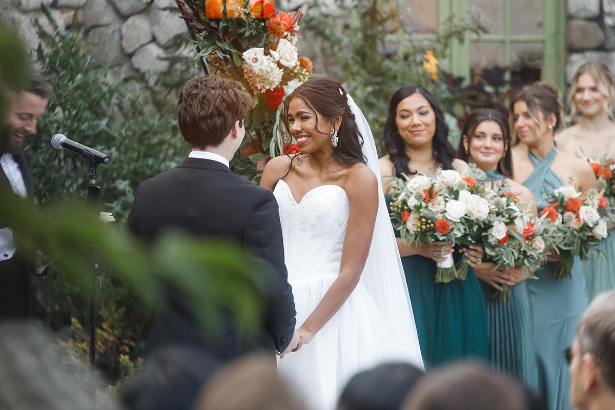 Bride and groom smiling at altar with bridesmaids holding bouquets in background.