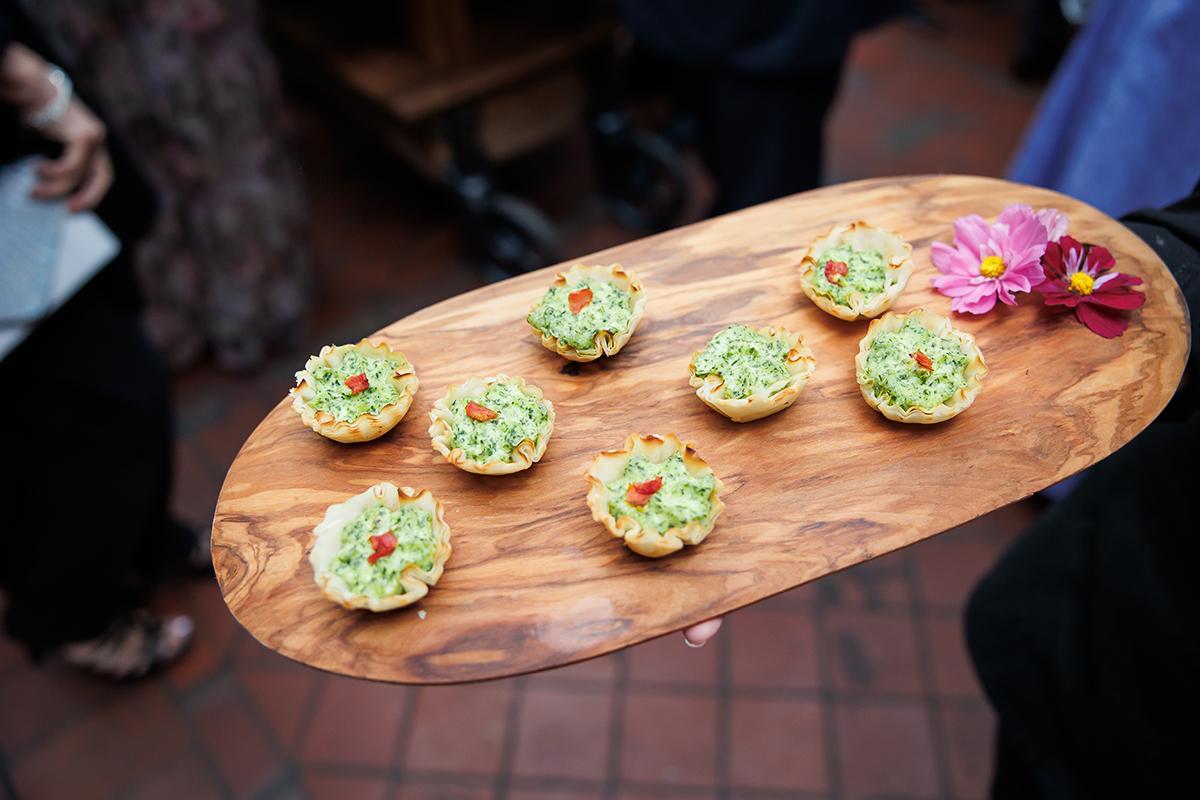 Tartlets with green topping on a wooden tray, next to pink flowers.