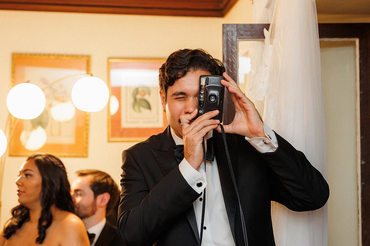 Man in a tuxedo using an instant camera at an indoor event.