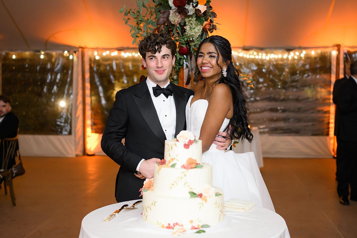 Wedding couple smiling beside a floral cake, dressed elegantly in a tented venue.