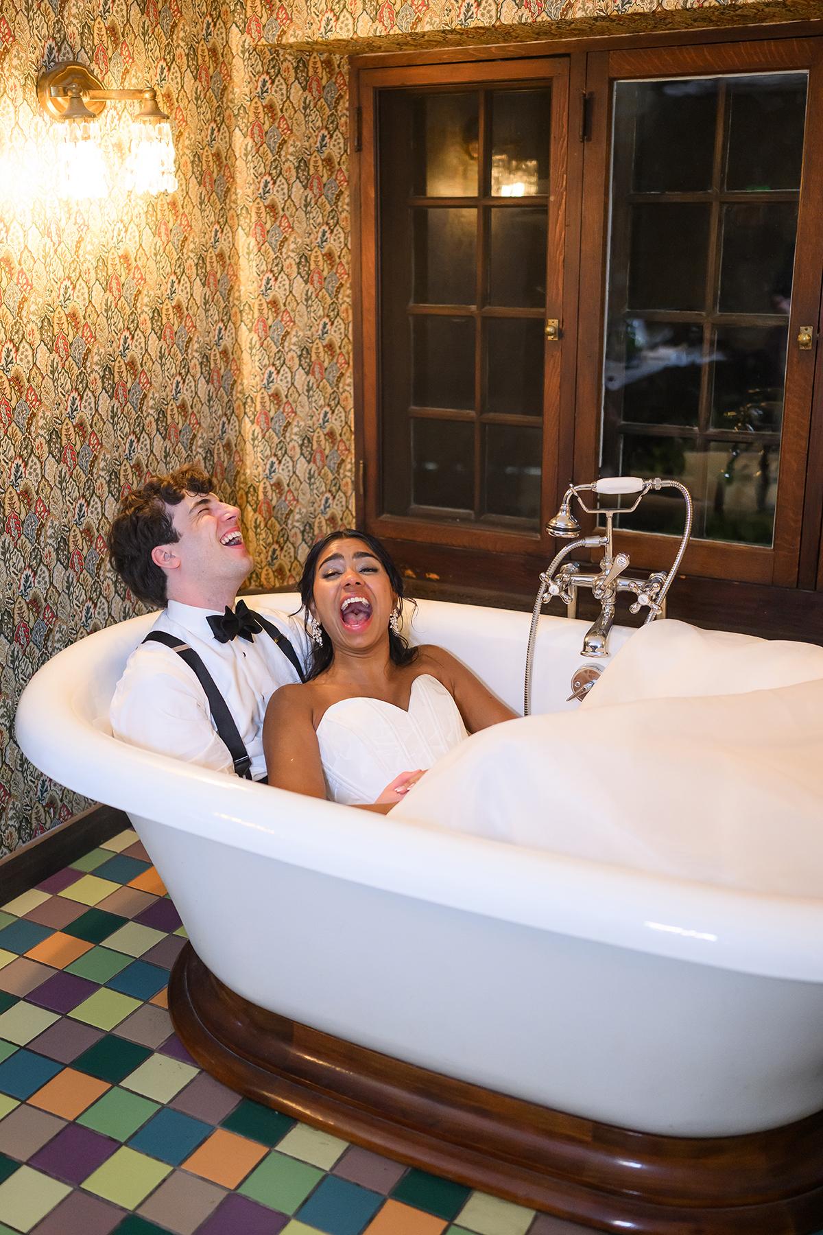 Couple laughing in a vintage clawfoot tub with floral wallpaper background.