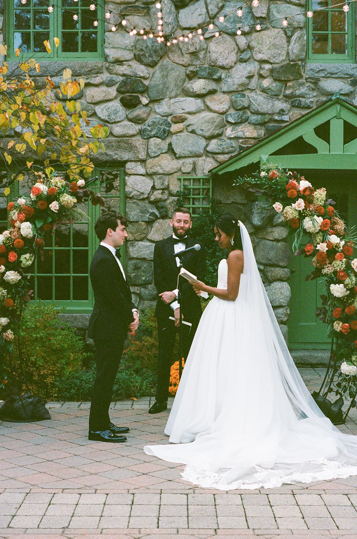 Bride and groom exchange vows outdoors next to stone wall and floral arch.