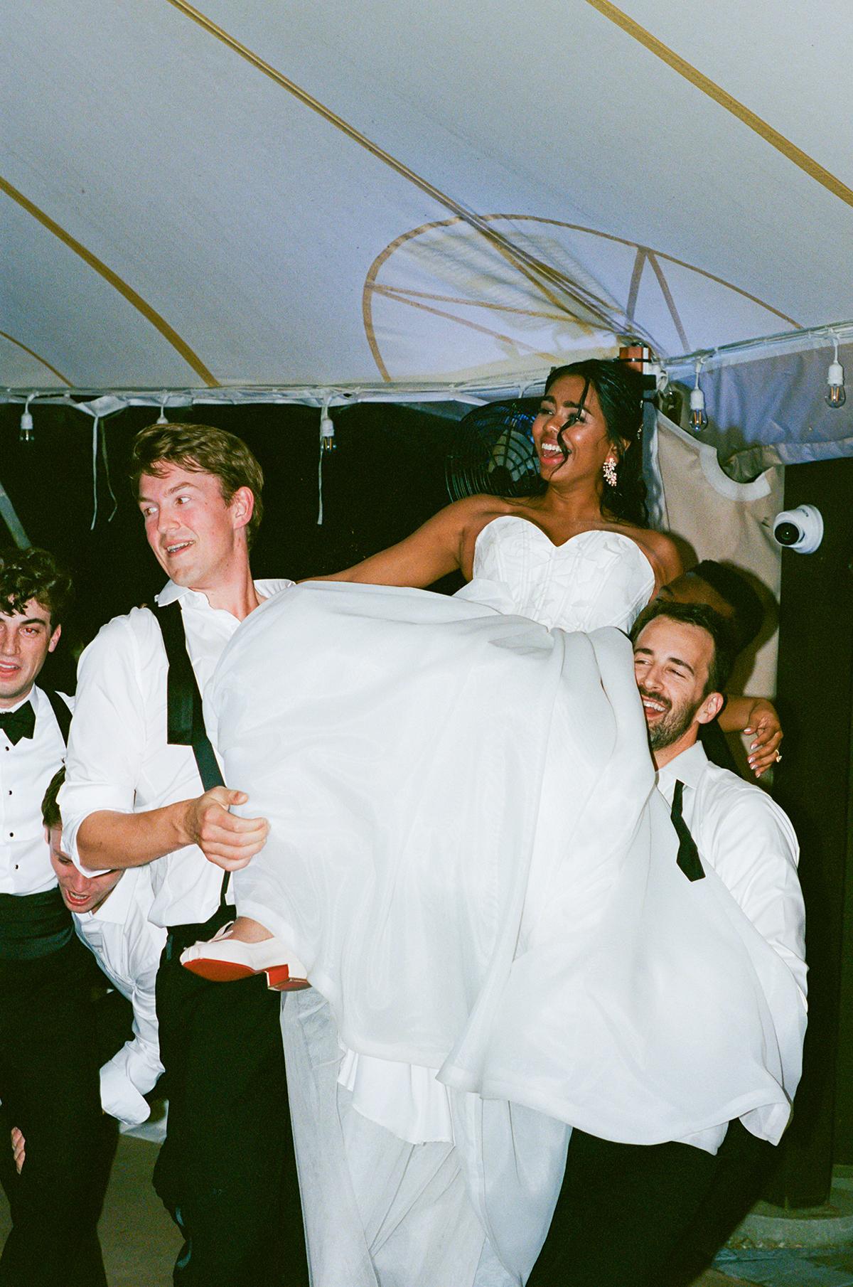 Bride in white dress is joyfully lifted by three men at a wedding reception.