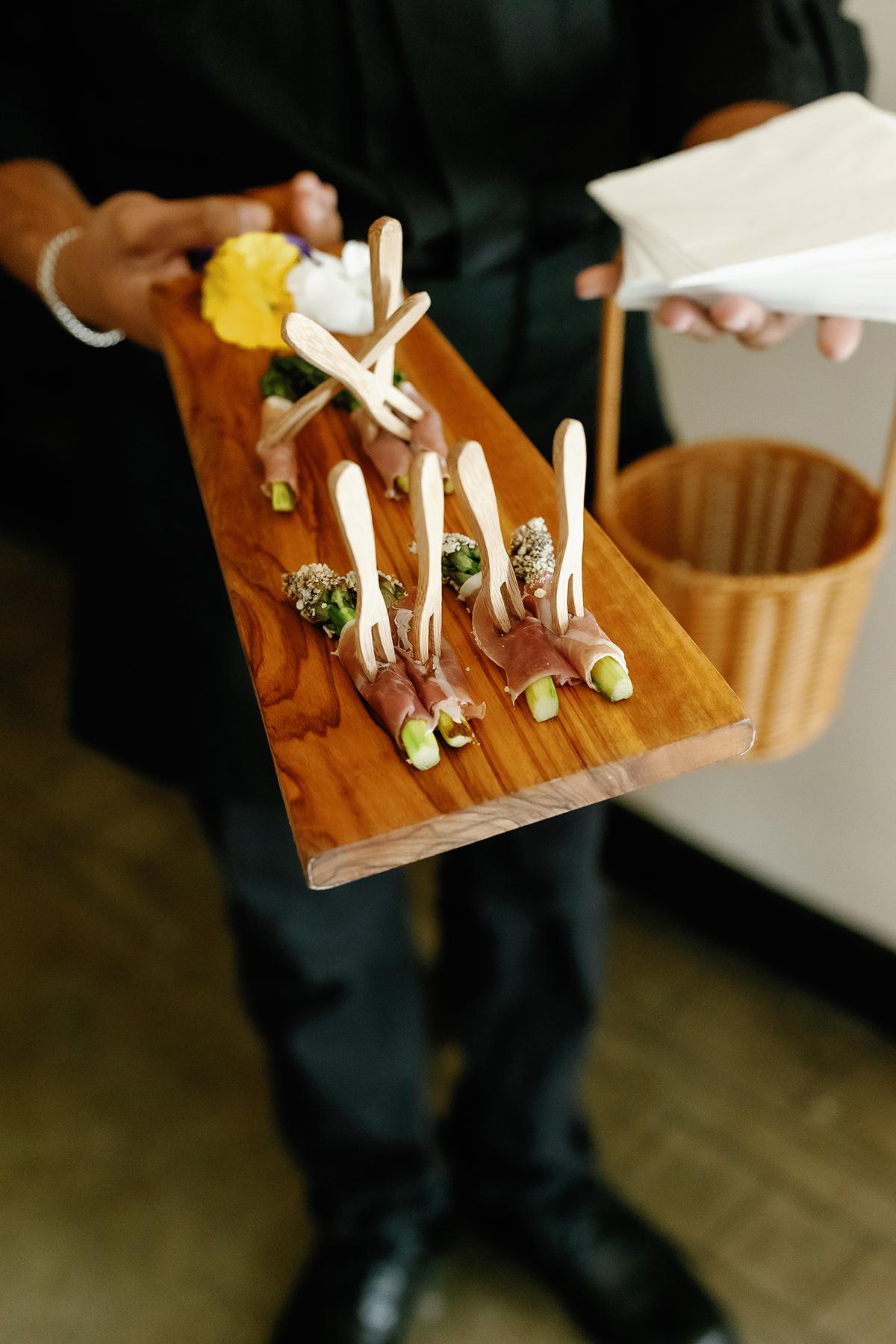 Server holding a tray of appetizers with asparagus and meat on wooden skewers.
