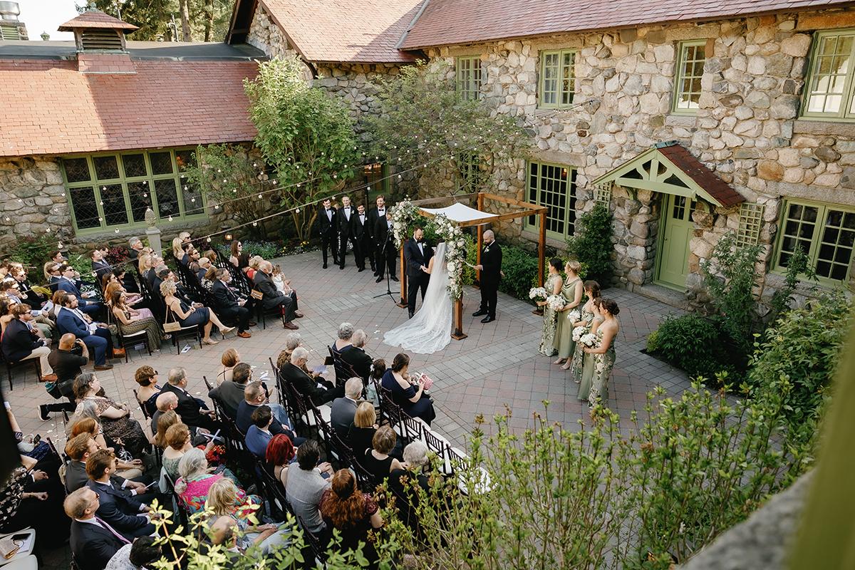 Outdoor wedding ceremony in stone courtyard, guests seated, couple under floral arch.