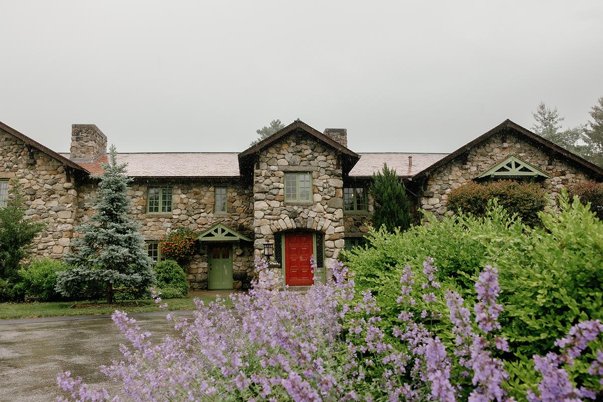 Stone house with red door and lush garden, purple flowers in foreground.