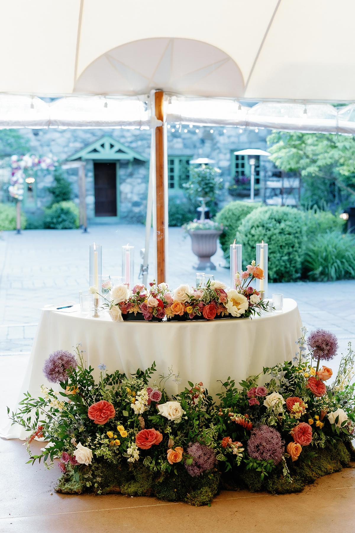 Round table with colorful floral centerpiece under a tent, garden background.