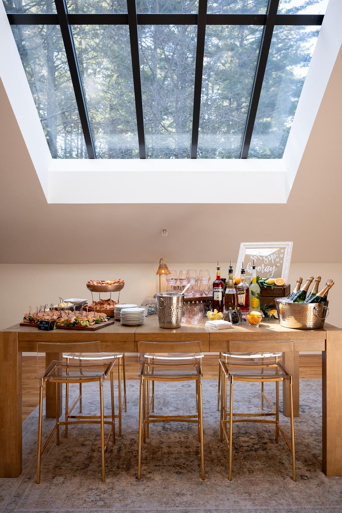 Elegant dining setup under a skylight with drinks and food on a wooden table.