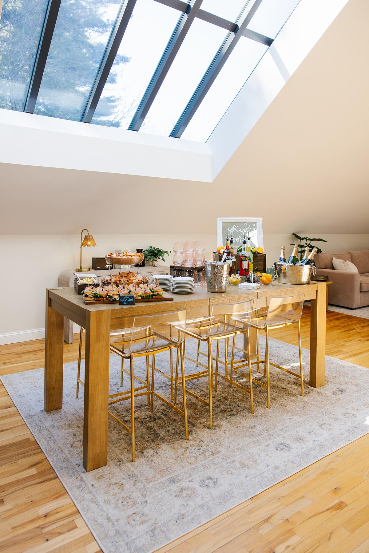 Modern dining area under skylight, with wooden table, stools, and assorted snacks.