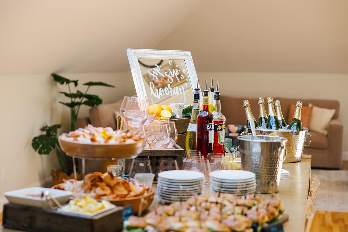 Buffet table with various appetizers, champagne bottles, and plants in a cozy room.