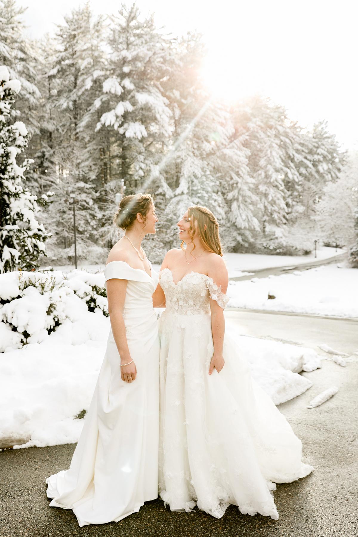 Two brides in white dresses stand in a snowy landscape, sunlight through trees.